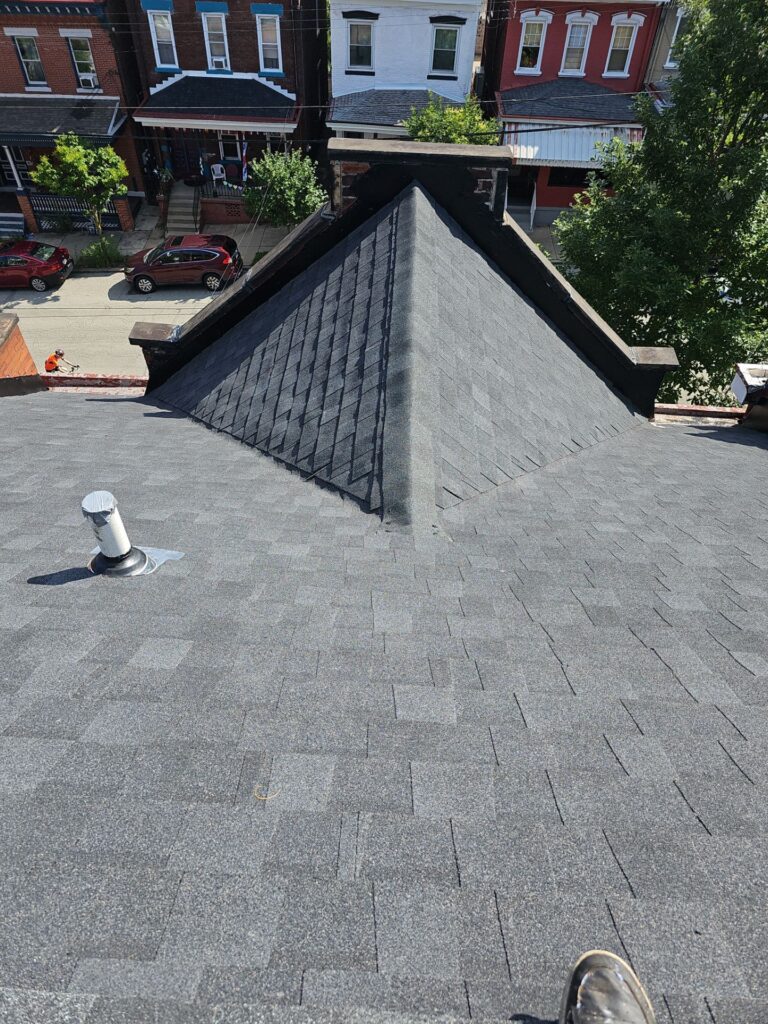 Roof view showcasing freshly installed dark shingles in Lawrenceville, Pittsburgh, with residential buildings and greenery in the background.