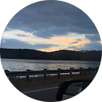 Scenic view of a lake at dusk, with dark clouds and a hint of sunset reflecting on the water, framed by a roadside barrier.