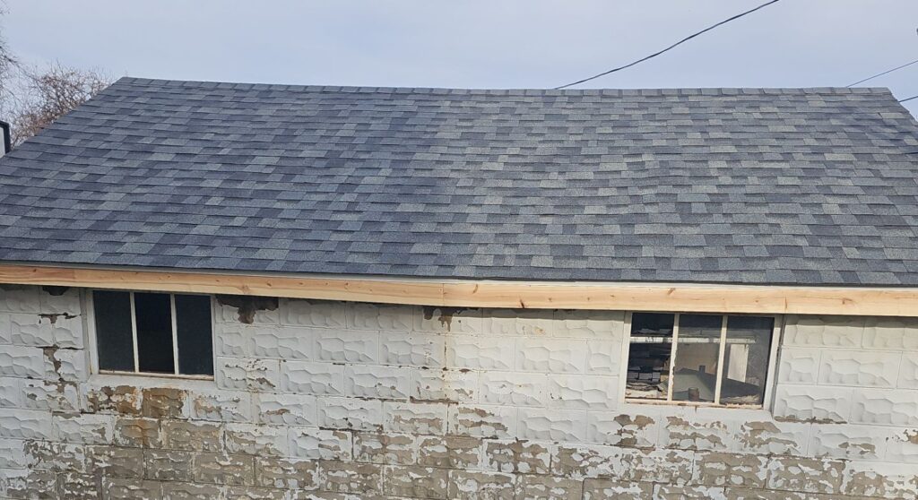 Newly installed asphalt shingle roof on a garage, showcasing quality workmanship and premium materials, with visible windows and stone wall below.