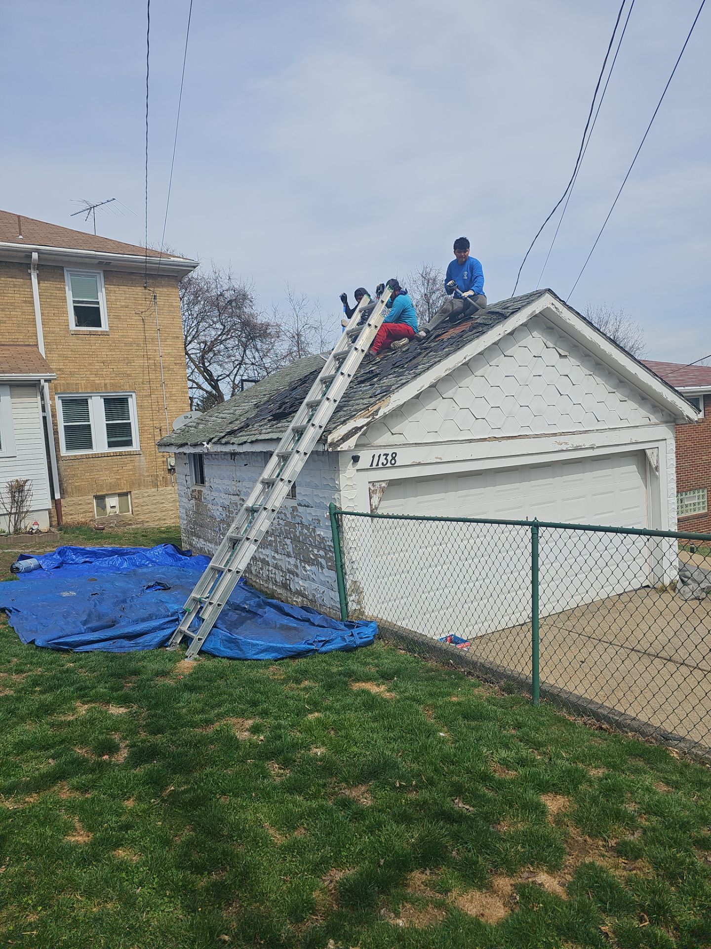 Workers replacing shingles on a garage roof, using a ladder and tarps for debris management, in a residential Beechview neighborhood.