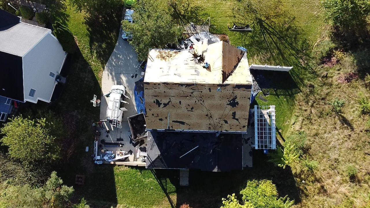 Aerial view of a house undergoing complete roof replacement, showing removed shingles and exposed roof deck, with surrounding debris and equipment visible, illustrating the roofing process by Alan Construction in Mount Washington.