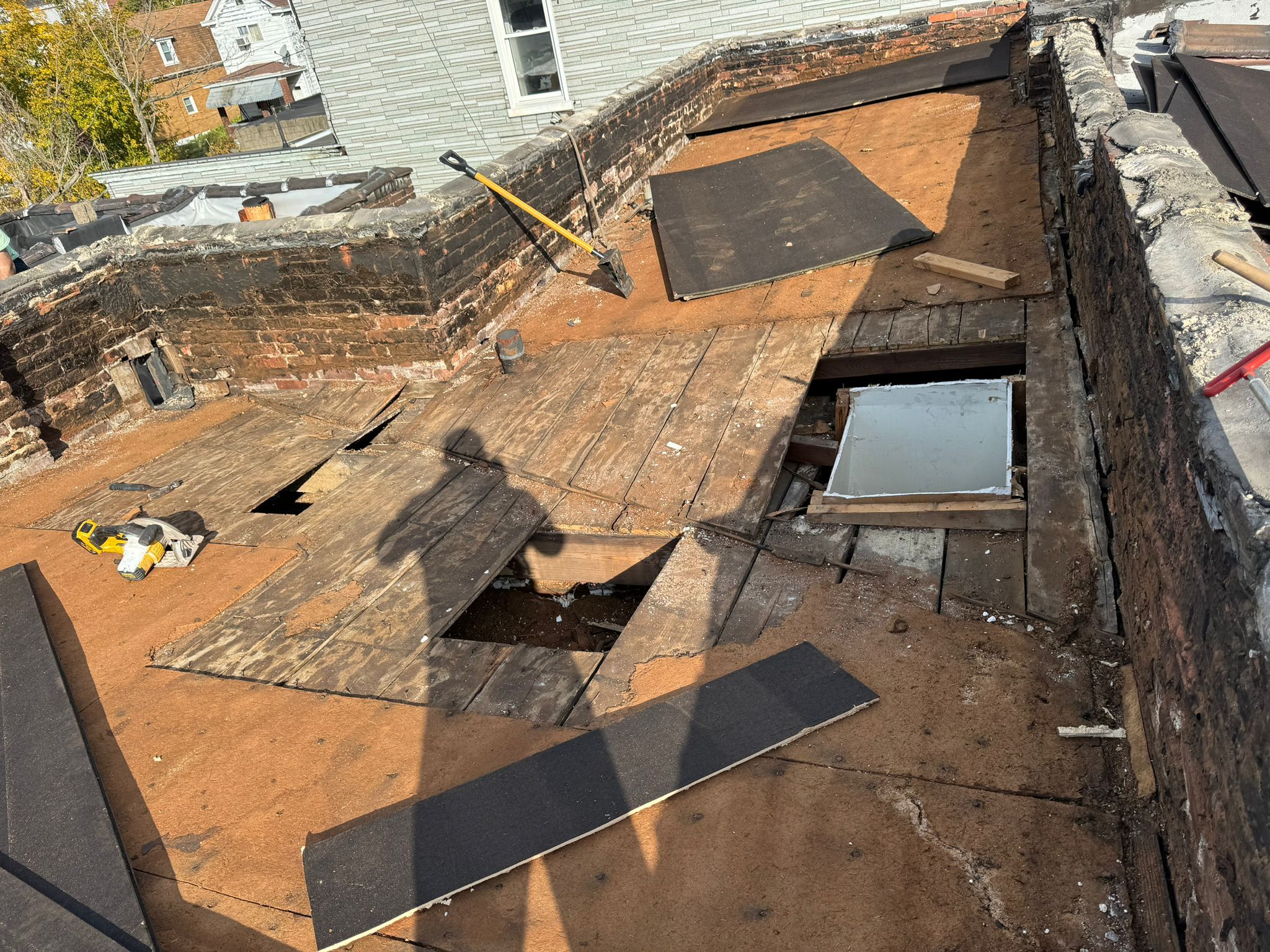 Flat roof deck undergoing preparation for EPDM rubber roofing installation, with removed wooden planks and visible repairs, tools scattered, and a shadow of the worker.