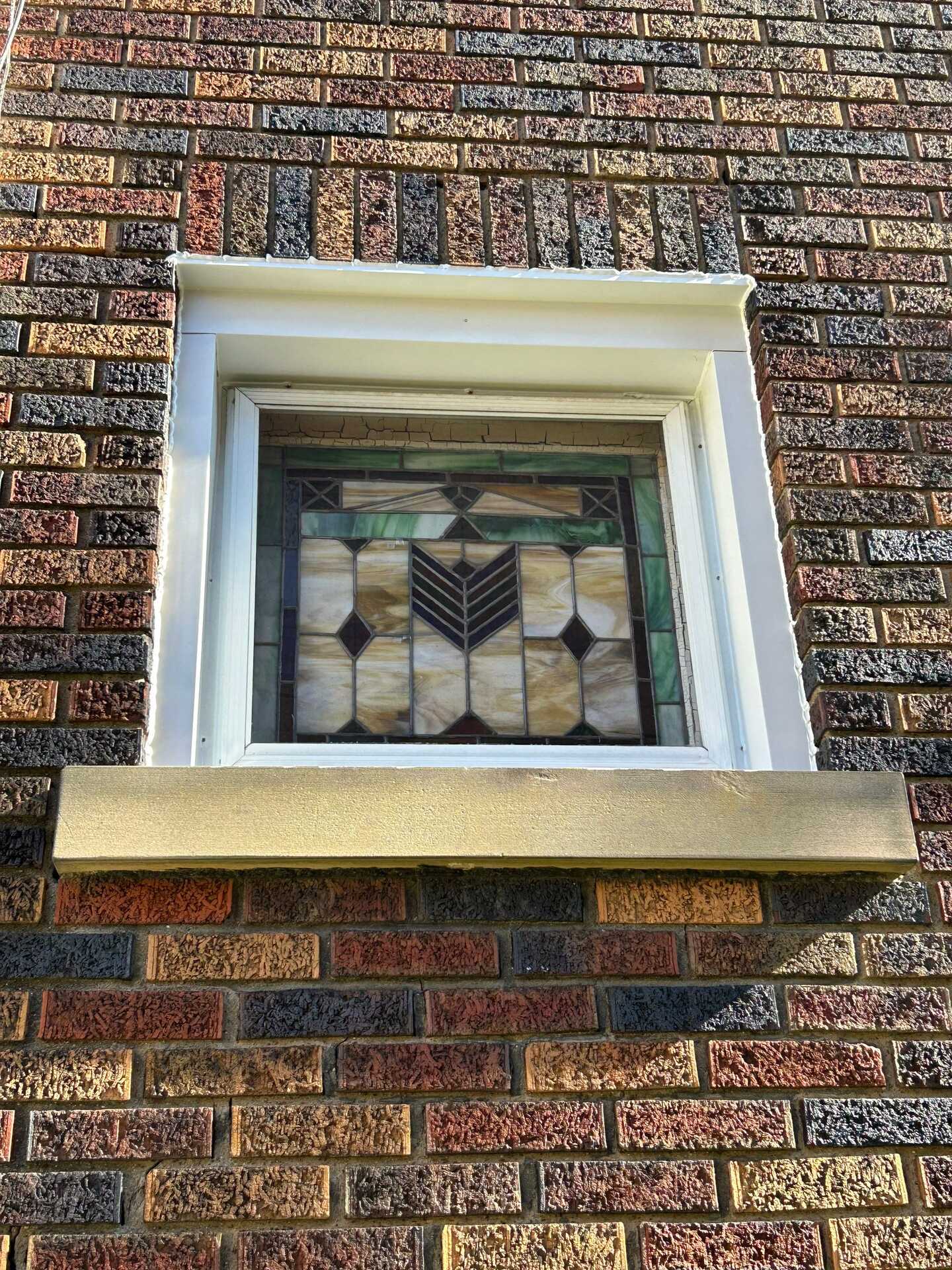 Stained glass window framed in white trim on a brick exterior, showcasing intricate geometric patterns, part of a home in Perry North undergoing window flashing repair.