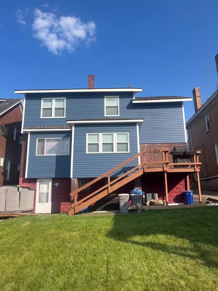 Rear view of a blue-sided two-story home with a wooden deck and green lawn.