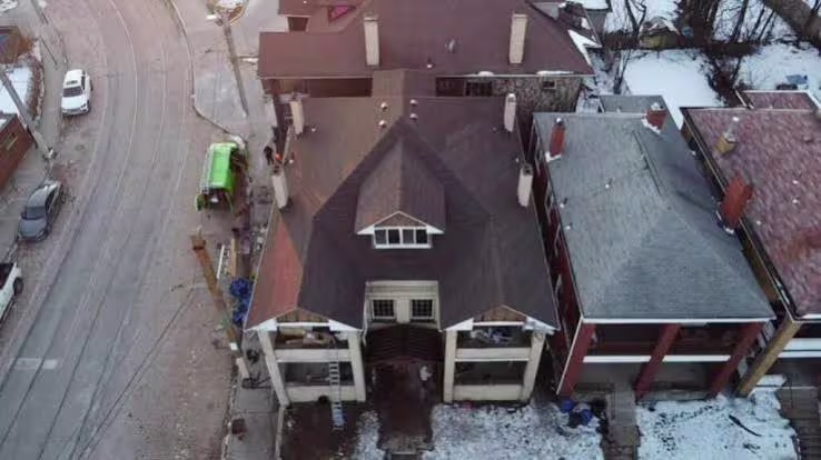 Aerial view of a snow-covered residential building with a steep roof and surrounding city street.