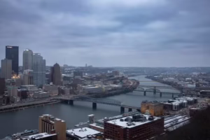 Winter skyline view of downtown Pittsburgh with snow-covered buildings, rivers, and bridges.