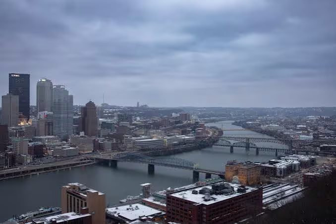 Winter skyline view of downtown Pittsburgh with snow-covered buildings, rivers, and bridges.