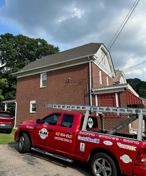 Red truck with "Alan Construction" branding parked outside a residential building, showcasing recently completed roofing work, featuring a shingled roof and brick exterior, emphasizing roofing, siding, and window services in Pittsburgh.